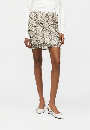 Kobieta w beżowej mini spódnicy z czarną kwiatową koronką, białym długim rękawem i białych szpilkach z małą kokardką.