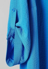 Blå strukturerad tygärm uppkavlad med en blå knappstängning mot en ljusgrå bakgrund.