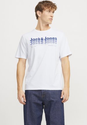 Young man wearing white short-sleeve t-shirt with blue "Jack & Jones" text and dark blue jeans, looking to his right against plain background.