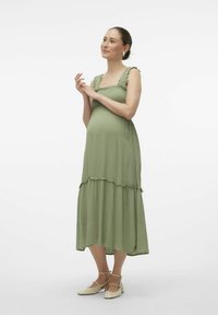 Vestido de maternidad en verde claro, con tirantes con volantes, diseño en capas y una silueta fluida. Llevado con zapatos planos en un amarillo pálido.