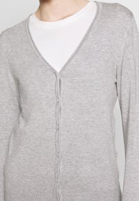 Ljusgrå cardigan med V-halsdesign, knäppning fram och ribbade kanter; tillverkad av ett mjukt, texturerat tyg.