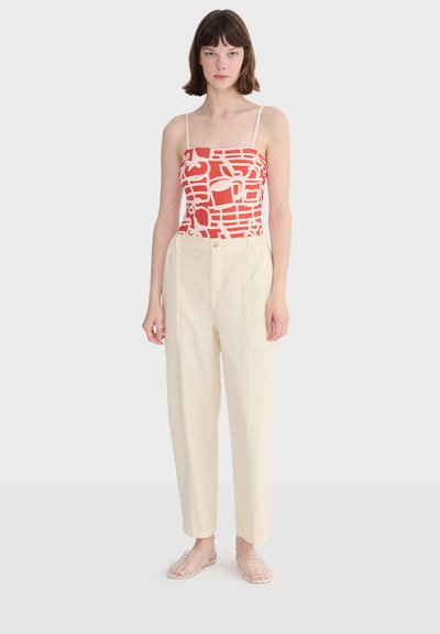 Jeune femme portant un débardeur à imprimé abstrait rouge et blanc, un pantalon crème taille haute et des chaussures transparentes à enfiler, debout sur un fond blanc.