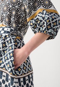 Bras d'une femme dans une robe à motifs avec des dessins géométriques noirs, blancs, jaunes et verts, main placée dans la poche latérale.