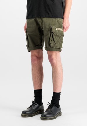 Homme portant un short cargo vert olive avec des poches latérales, des chaussettes noires courtes et des chaussures noires à lacets sur fond blanc.