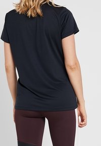 Kvinde med skulderlangt hår iført en sort T-shirt med korte ærmer og bordeauxfarvede leggins med sidelister, set fra ryggen.