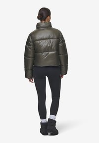 Korte, puffy olijfgroene jas met een hoge kraag en glanzende afwerking, gecombineerd met zwarte leggings en laarzen. Gestructureerd en gewatteerd ontwerp.