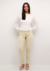 Hvid, rynket bluse med lange ærmer og flæsekant, kombineret med lyse beige, tætsiddende bukser og nøgenfarvede højhælede sko. Simpelt design, glat stof.