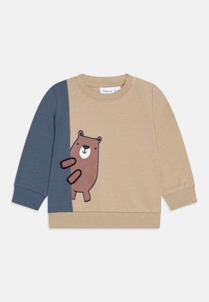 Felpa beige con manica blu, caratterizzata da un'illustrazione fantasiosa di un orso marrone sul davanti. Tessuto morbido e polsini a costine.