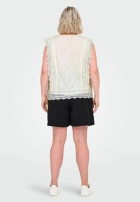 Kvinne med kort blondt hår iført en ærmeløs kremfarget blondebluse, sorte shorts og hvite joggesko, står med ryggen til på en ensfarvet bakgrunn.