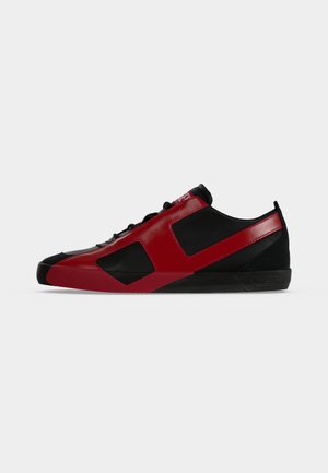 Zapatilla baja en negro y rojo con cierre de cordones y diseño de panel geométrico sobre fondo blanco.