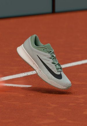 Svetlo zelena tekaška superga Nike s črnim logotipom, ki je v zraku, brca rdečo glino in bele črte igrišča v ozadju.