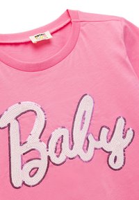 Vaaleanpunainen puuvillapaita, jossa on "Baby" -teksti strassikirjailla. Pyöreä kaula-auko ja lyhyet hihat; pehmeä kuitu kiiltävillä yksityiskohdilla.