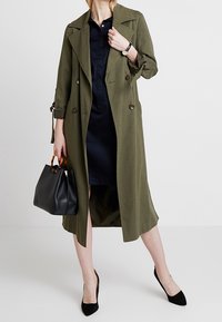 Donna che indossa un trench coat verde oliva sopra un vestito nero, tiene una borsa nera con manici in tartaruga, e delle scarpe con tacco nere.