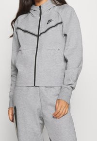 Grå zip-up jacka i mjukt tyg, med hög krage, svarta detaljer och ett chevron-mönster på bröstet. Inkluderar två sidfickor.