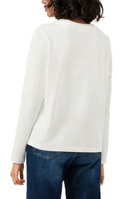 Biała bawełniana koszula z długim rękawem, okrągłym dekoltem i luźnym krojem. Posiada boczne rozcięcia; zestawiona z niebieskimi dżinsami.