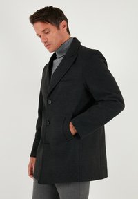 Mustast villast mantlit, millel on struktuurne lõige, sälkrutud krae, kaks nööpi, küljetaskud ja rinnatasku; kantud halli kaelusega pulloveri peal.