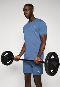 Camiseta deportiva azul con logo en el pecho, combinada con pantalones cortos a juego. El modelo sostiene una barra de pesas negra con discos redondos, mostrando brazos musculosos.