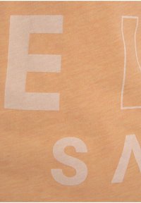 Perzikgekleurde stof met een zachte textuur, voorzien van grote witte letters. Het ontwerp bevat abstracte vormen en variërende letterhoogtes.