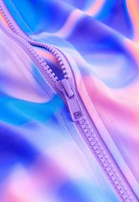 Detailný záber na zips levandulovej farby, čiastočne zapnutý na hladkej látke s pastelovým prechodom modrej, ružovej a fialovej farby.