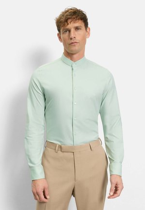 Mann trägt ein hellgrünes Hemd mit Stehkragen und beige Hose, steht vor einem einfarbigen hellen Hintergrund.