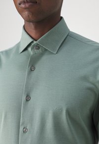 Myntgrön polo skjorta med en texturerad tyg, knappad krage och grå knappar. Har en diskret broderad logotyp på bröstet.