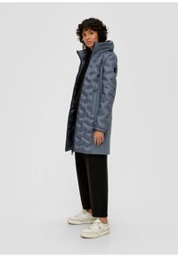 Blauer gepolsterter Mantel mit hohem Kragen, ausgestattet mit einem Reißverschluss, Seitentaschen und einer glatten Oberfläche. Kombiniert mit schwarzen Hosen und hellen Turnschuhen.