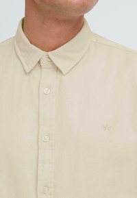Camisa beige de botones de algodón, con cuello, botones frontales y un pequeño logo bordado en el pecho. Textura suave.