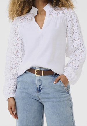 Kvinde iført hvid bluse med blondærmer og lyseblå jeans, stylet med brunt bælte og hånd i lommen.