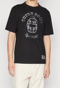 Il modello indossa una maglietta nera con una grafica di un casco da football e la scritta "SUPER BOWL LVIII Las Vegas", abbinata a pantaloni chiari.