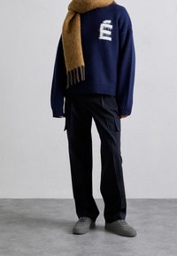 Navy knitted sweater with white logo, paired with black cargo trousers and gray sneakers. Accented by a long, tan fringed scarf.