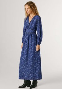 Mėlyna ilga suknelė su gėlių raštu, V formos iškirpte, ilgomis rankovėmis ir elastine liemens juosta. Derinama su juodais aulinukais su kulnais.