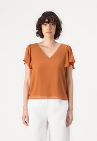Top de manga corta con escote en V en color óxido, hecho de tela ligera con adornos de volantes en las mangas, combinado con pantalones blancos.