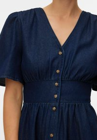 Nærbilde av en person iført en mørkeblå denimkjole med korte, puffede ermer og en rekke brune knapper ned foran.