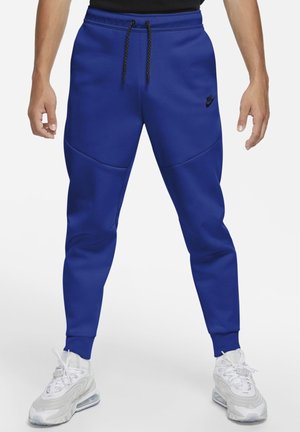 Pantalon de jogging bleu Nike avec cordon noir, logo noir sur la cuisse gauche, porté avec des chaussures de sport blanches sur une personne debout.