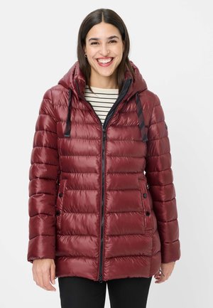 Smiling woman wearing a maroon quilted hooded jacket over a striped shirt and black pants against a plain light background.