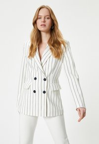 Blazer blanco de doble botonadura con rayas verticales marinas, que presenta botones negros, solapas de pico y hombros estructurados. Textura suave.