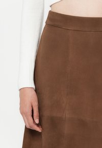 Jupe en daim marron avec une texture lisse, présentant une taille cintrée et des coutures subtiles. Manche d'un pull en tricot blanc partiellement visible.