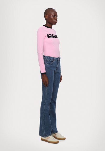 Pink long-sleeve shirt with black neckline and graphic text, paired with blue flared jeans and beige shoes with multicolored stripes.
