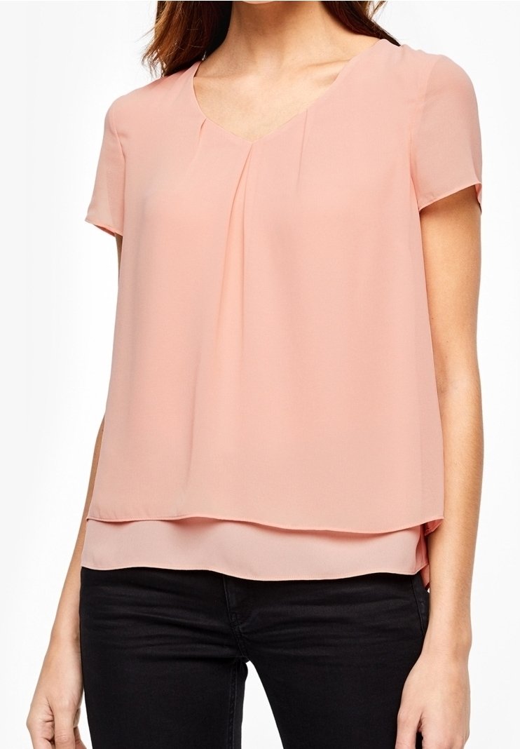 Femme portant un chemisier à manches courtes rose clair à superpositions, associé à un pantalon noir, visible des épaules aux hanches sur un fond blanc.