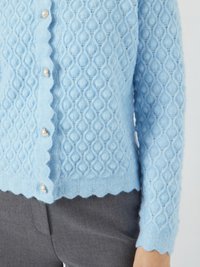 Gilet en tricot bleu clair avec un motif de losanges texturé, un ourlet festonné et des boutons ressemblant à des perles sur le devant. Porté sur un pantalon gris.