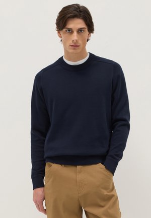 Giovane uomo con capelli castani indossa un maglione blu navy a girocollo sopra una camicia bianca e pantaloni kaki, in piedi contro uno sfondo chiaro e uniforme.