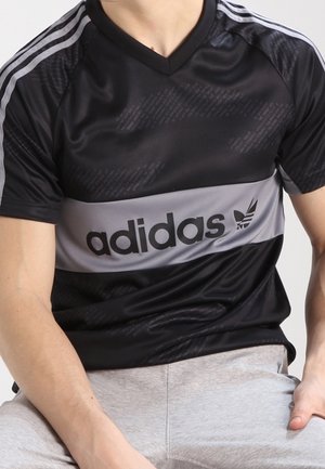 Man draagt een zwart-wit Adidas sportshirt met logo op de borst, gecombineerd met lichtgrijze broek, zittend tegen een effen achtergrond.