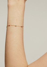 Pulsera de cadena de oro con tres piedras preciosas de colores (rojo, verde, azul) colocadas a lo largo de la cadena. Llevada en un brazo de piel clara.