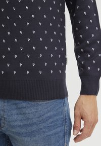 Navy gestrickter Pullover mit sich wiederholendem weißen Herzmuster, gerippten Bündchen und Saum. Kombiniert mit hellblauen Jeans.