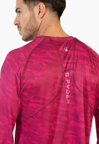 Vaaleanpunainen pitkähihainen urheilupaita, jossa on teksturoitu kuosi, litteät saumat ja pieni hämähäkkilogo selässä kaula-aukon lähellä.