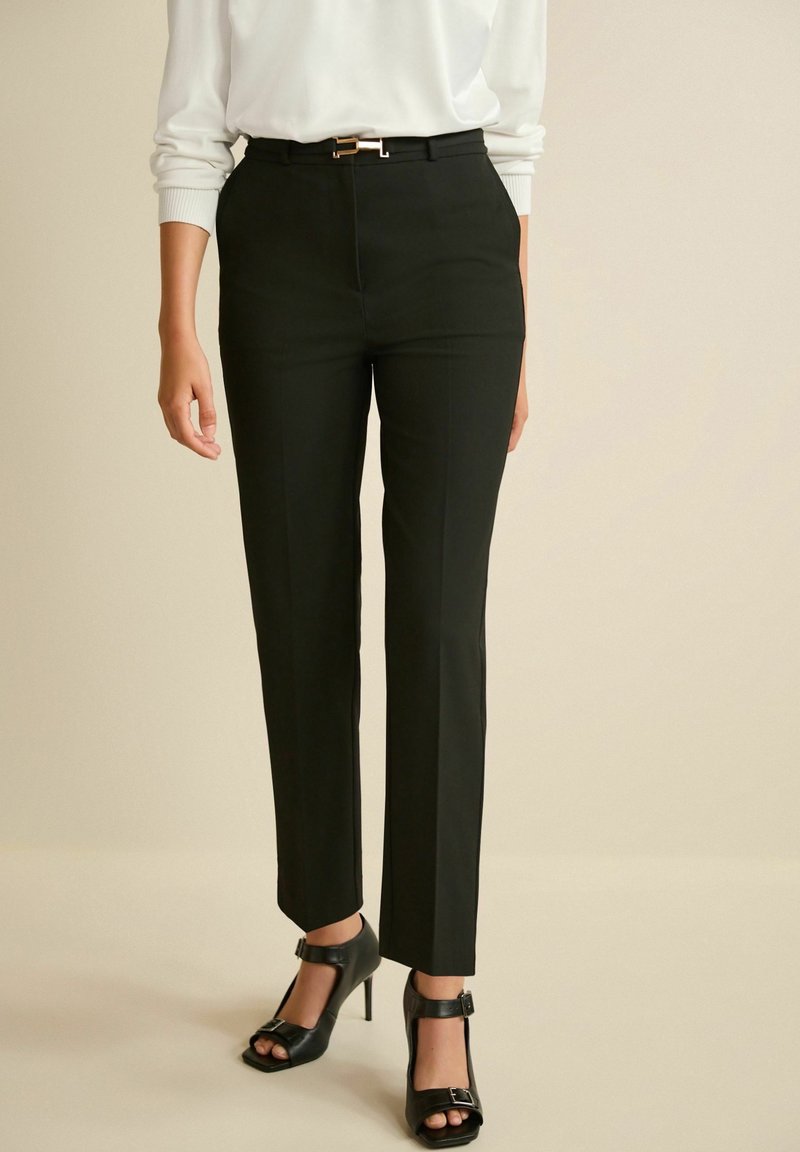 Femme portant un pantalon noir sur mesure avec ceinture à l'avant et des chaussures noires à talons hauts ouvertes à l'avant, debout devant un fond uni.
