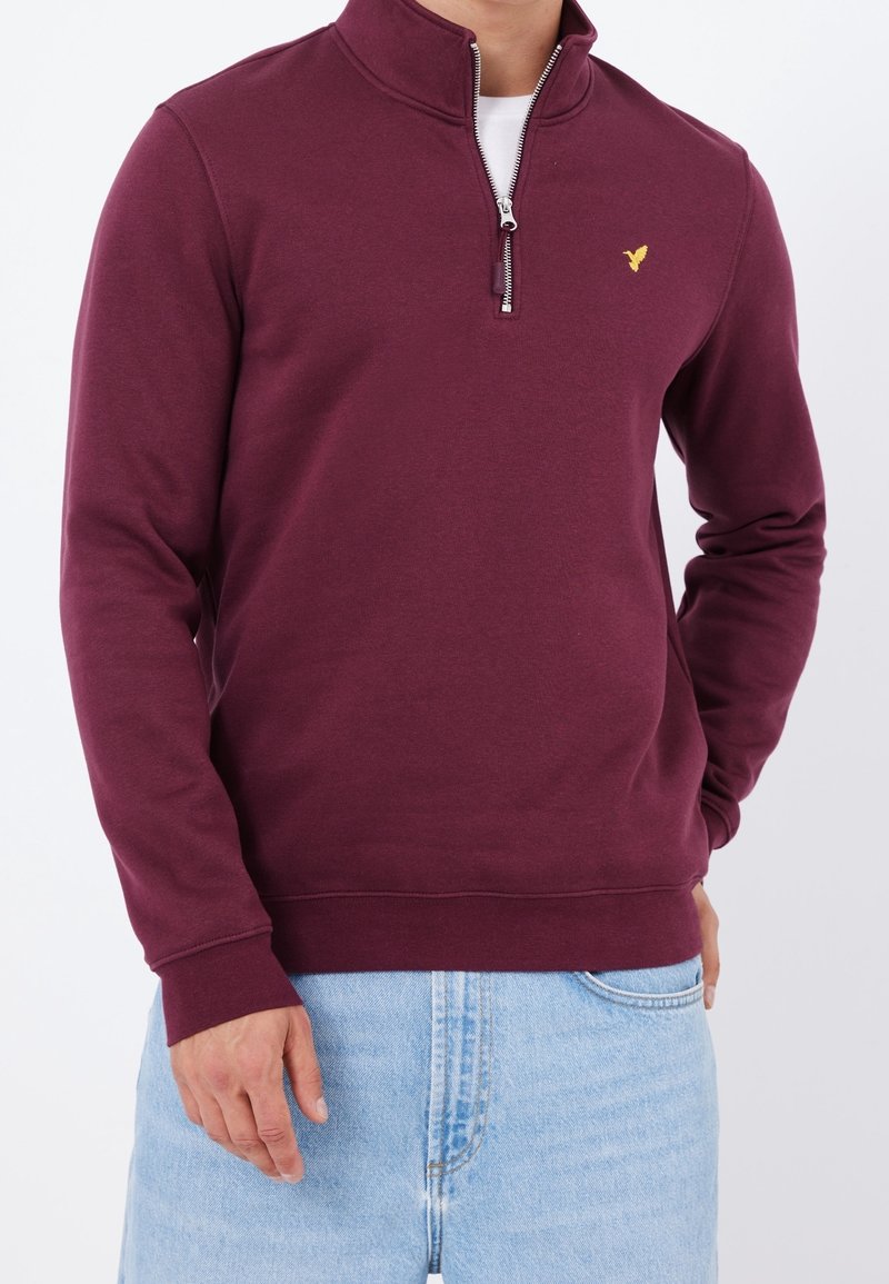 Burgundská mikina na zip s vysokým límcem, s malým žlutým logem na levé straně hrudi a žebrovanými lemovkami na rukávech a spodním okraji.