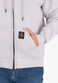 Lichtgrijze zip-up hoodie met een voorvak, capuchon met trekkoord en een klein zwart logo-opstiksel. Beschikt over geribde manchetten en onderrand.