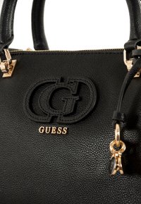 Sort faux læder håndtaske med en struktureret overflade, præget logo, guld hardware og en dekorativ charm fastgjort til håndtaget.