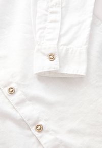 Poignet et devant d'une chemise blanche à manches longues avec des boutons beiges sur un fond en tissu texturé.